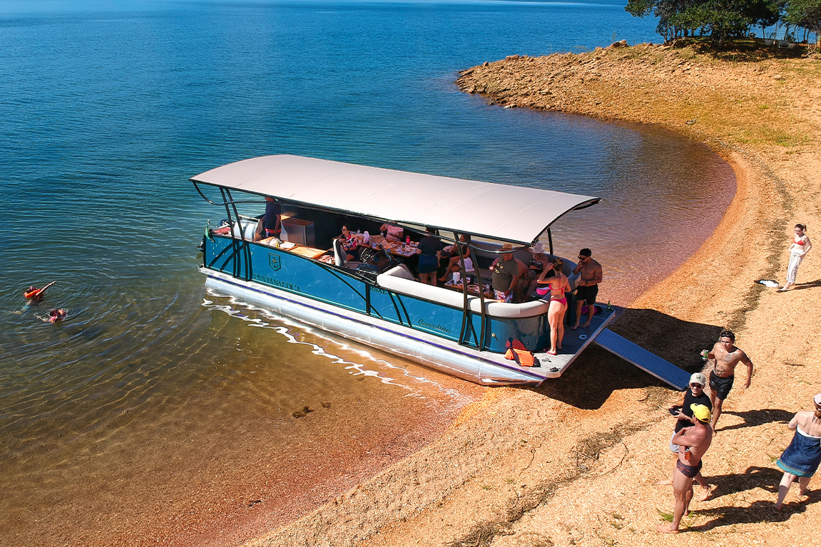 Pontoon R5 SXL atracado de ré em uma praia de água doce, com pessoas descendo pela plataforma para aproveitar o dia de sol e a natureza.